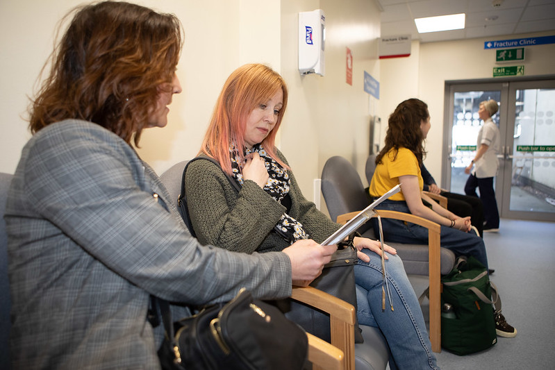 Two women sit in a hospital waiting area, one with a tablet and the other looking on. Two more people are seated further down the row of chairs, and a healthcare worker is visible walking in the background. Signs on the wall indicate that the area is for a Fracture Clinic.