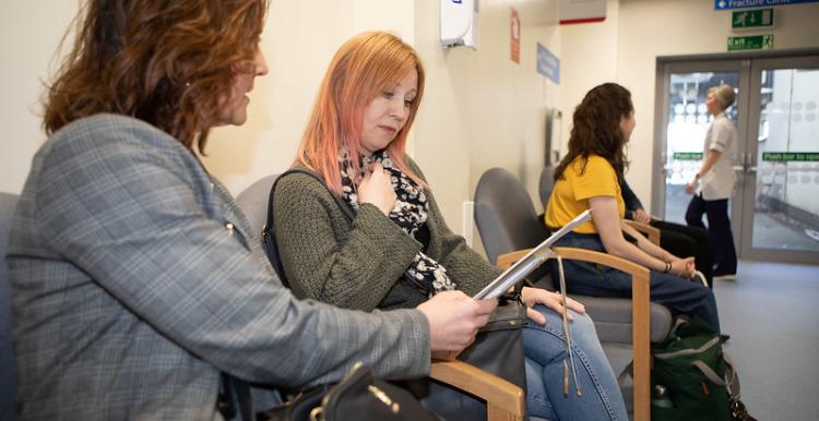 Two women sit in a hospital waiting area, one with a tablet and the other looking on. Two more people are seated further down the row of chairs, and a healthcare worker is visible walking in the background. Signs on the wall indicate that the area is for a Fracture Clinic.