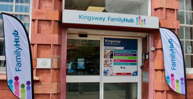 Entrance to Kingsway Family Hub in Widnes, showing a red-brick building with a sign above the door reading "Kingsway Family Hub" and two banners on either side displaying the Family Hub logo.