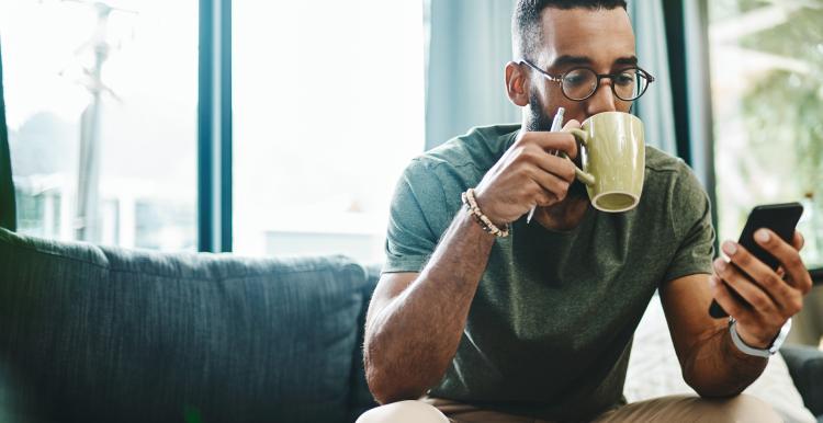 A man uses his mobile phone whilst drinking from a mug. 