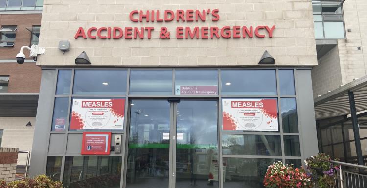 Entrance to Whiston Hospital Children’s Accident & Emergency department, showing glass double doors beneath red “Children’s Accident & Emergency” signage. Measles information posters are displayed on both sides of the entrance, with flower planters positioned near the doorway.
