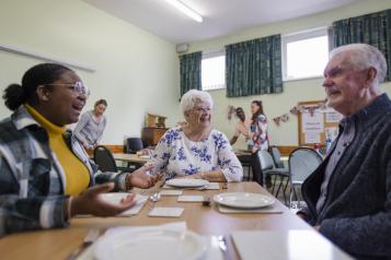 People chat, at a dinner table, in a community centre. 
