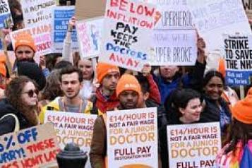 Image of Junior Doctors holding up protest signs on a picket line. Signs read '£14/ hour is not a fair wage for a Junior Doctor full pay restoration now. BMA'