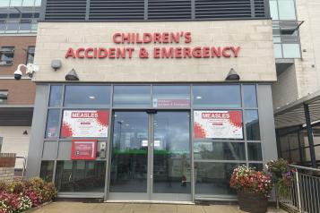 Entrance to Whiston Hospital Children’s Accident & Emergency department, showing glass double doors beneath red “Children’s Accident & Emergency” signage. Measles information posters are displayed on both sides of the entrance, with flower planters positioned near the doorway.