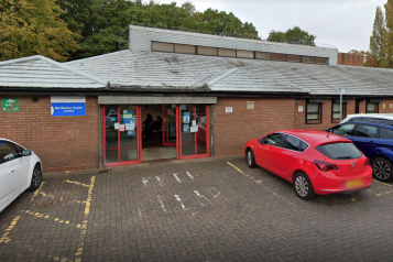 Exterior of Murdishaw Health Centre, a single-storey brick GP surgery with a sloped grey roof and red-framed glass entrance doors. Health centre signage is mounted on the brick wall to the left of the entrance. A small car park is directly in front, with marked bays and several parked cars. Trees are visible behind the building on an overcast day.