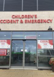 Entrance to Whiston Hospital Children’s Accident & Emergency department, showing glass double doors beneath red “Children’s Accident & Emergency” signage. Measles information posters are displayed on both sides of the entrance, with flower planters positioned near the doorway.